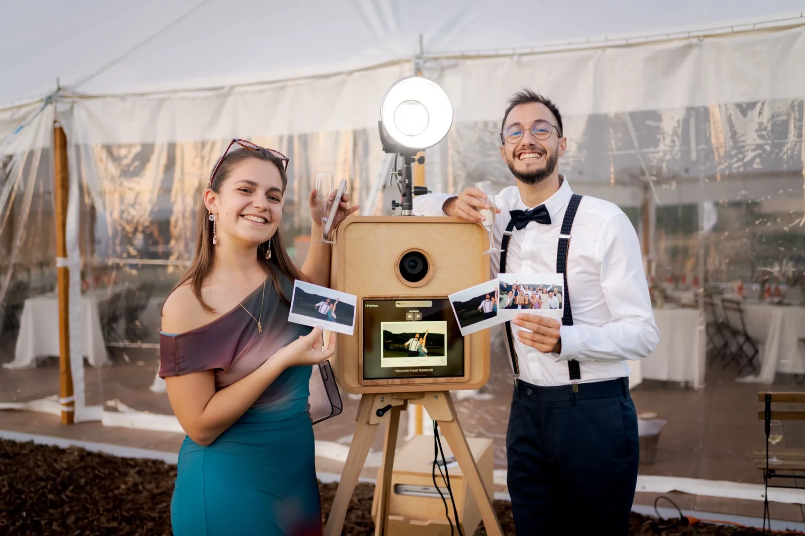 Deux invités souriants tenant leurs tirages photos devant le photobooth en bois MH lors d'un mariage, flash professionnel allumé